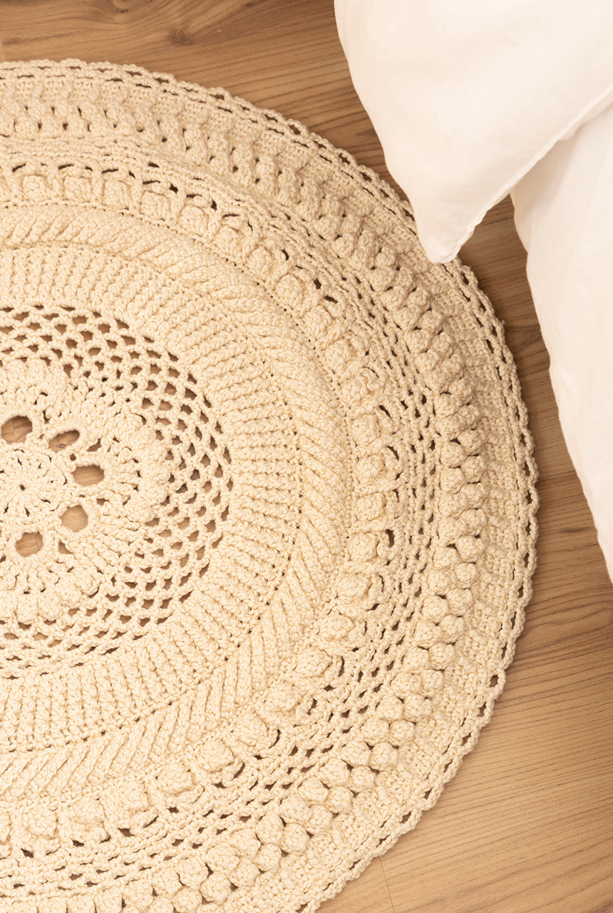 Circulo Barroco Natural Brilho - Natural Cotton Yarn Gold/Silver, 657m / 433m, 400g - A round, hand-crocheted area rug in a natural beige color, styled on a light wood floor next to a white pillow.