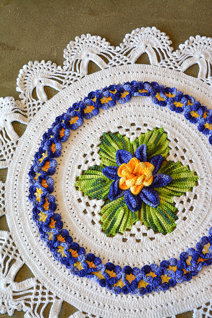 Circulo Barroco Multicolor Premium 4/6 - Brazilian Cotton Yarn, 226m/200g - A round crocheted doily with a white base, featuring a central green and blue flower motif with a yellow-orange center, surrounded by blue and yellow floral borders.