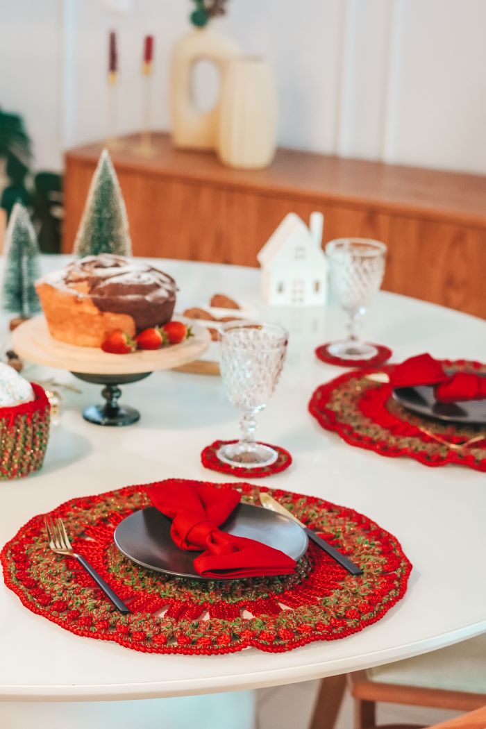 Círculo Barroco Maxcolor Sparkle – Cotton Yarn (216m / 200g) - A festive holiday table setting featuring red and gold crocheted placemats, dark plates, red napkins, and clear textured glasses, with a decorated cake and miniature Christmas trees in the background.