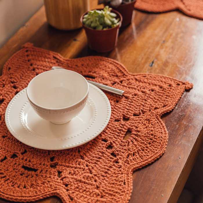 Círculo Barroco Maxcolor Sparkle – Cotton Yarn (216m / 200g) - An orange-brown crocheted table placemat with a scalloped edge, styled on a wooden table with a white ceramic bowl and saucer, a silver spoon, and small potted plants in the background.