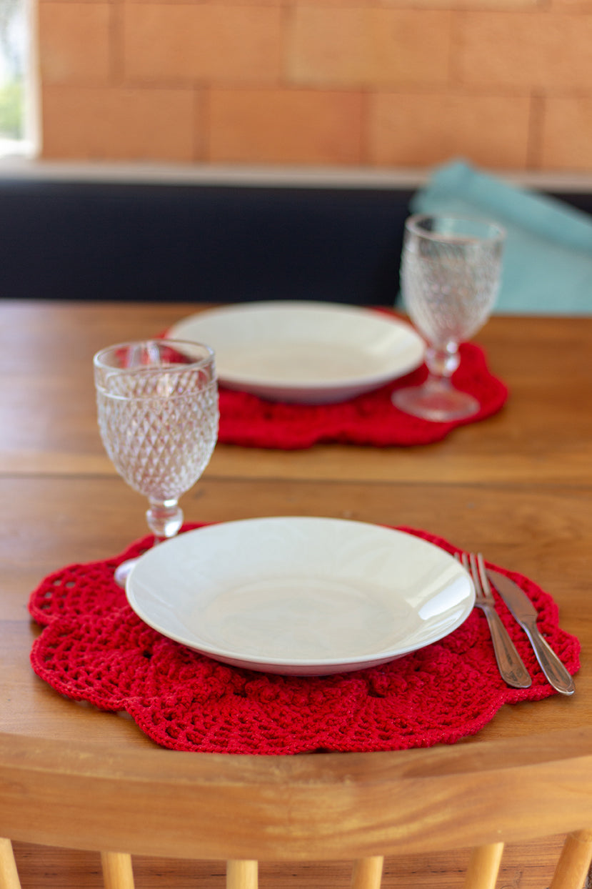 Círculo Barroco Maxcolor Sparkle – Cotton Yarn (216m / 200g) - Círculo Barroco Maxcolor Sparkle – Cotton Yarn (216m / 200g) - A close-up of a wooden dining table set with two white plates on red crocheted placemats, each plate accompanied by a clear cut-glass wine glass and silver cutlery, with a blurred indoor background.