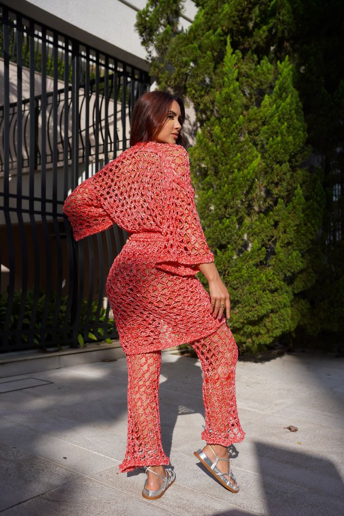 Circulo Jeans – 100% Brazilian Cotton Denim Effect Yarn (132m/100g) - A woman wearing a coral pink crochet knit two-piece set consisting of a loose long-sleeve top and matching wide-leg pants, standing outdoors on a paved walkway with green trees and a black metal fence in the background.
