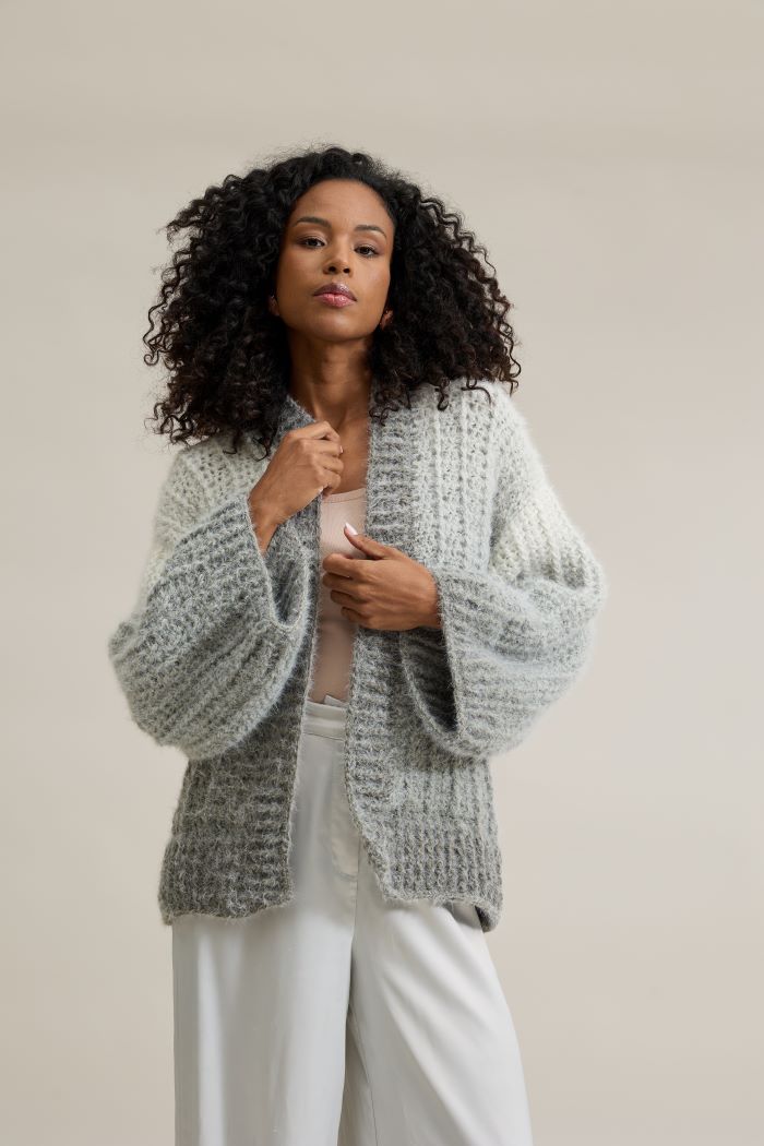 Círculo Marfim – Acrylic Gradient Cake Yarn (400 m / 200 g) - A woman with curly black hair wearing a chunky, textured light gray knit cardigan over a pale pink top and white pants, posing against a solid beige studio background.