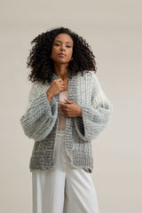 Círculo Marfim – Acrylic Gradient Cake Yarn (400 m / 200 g) - A woman with curly black hair wearing a chunky, textured light gray knit cardigan over a pale pink top and white pants, posing against a solid beige studio background.