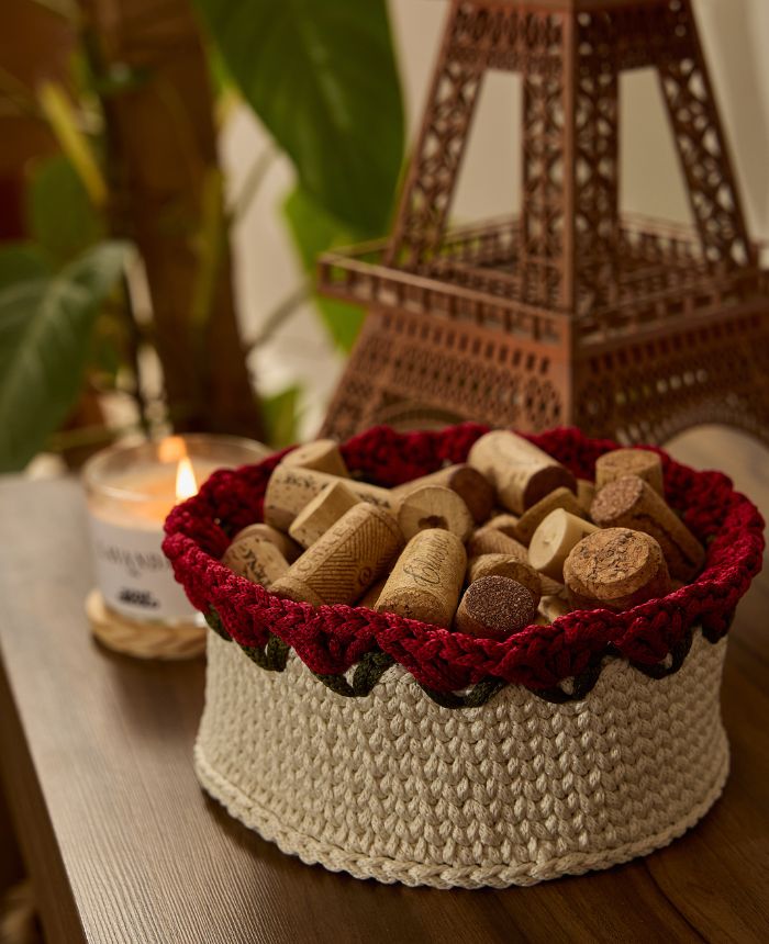 Circulo Fio Náutico Poly 5 mm – Polyester Cord Yarn ( 178 m  / 500 g) - A round crocheted basket with a cream-colored body and a deep red scalloped rim, filled with cork stoppers. The basket sits on a wooden table in a cozy setting with a lit candle and a decorative Eiffel Tower in the background.
