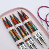 KnitPro Symfonie 2 Normal interchangeable needle set, 13 cm, Chunky - Six colorful wooden knitting needles with silver metal tips, secured in a pink zippered case with white elastic bands. Two purple cable knitting needles are coiled in the background on a white surface.